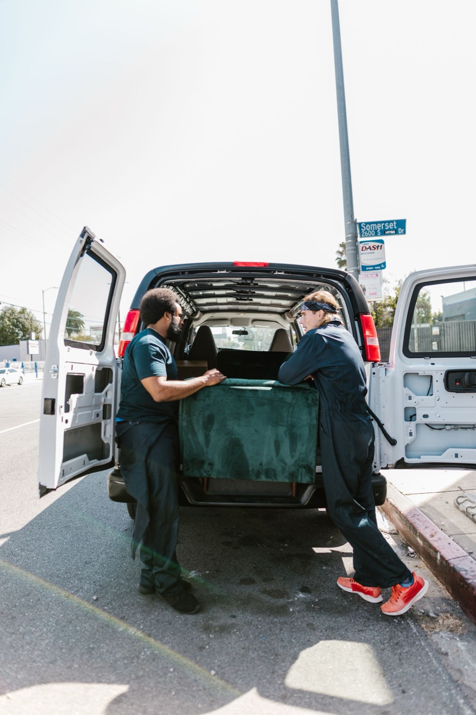 Two uniformed movers carefully loading a green sofa into the back of a moving van on a city street