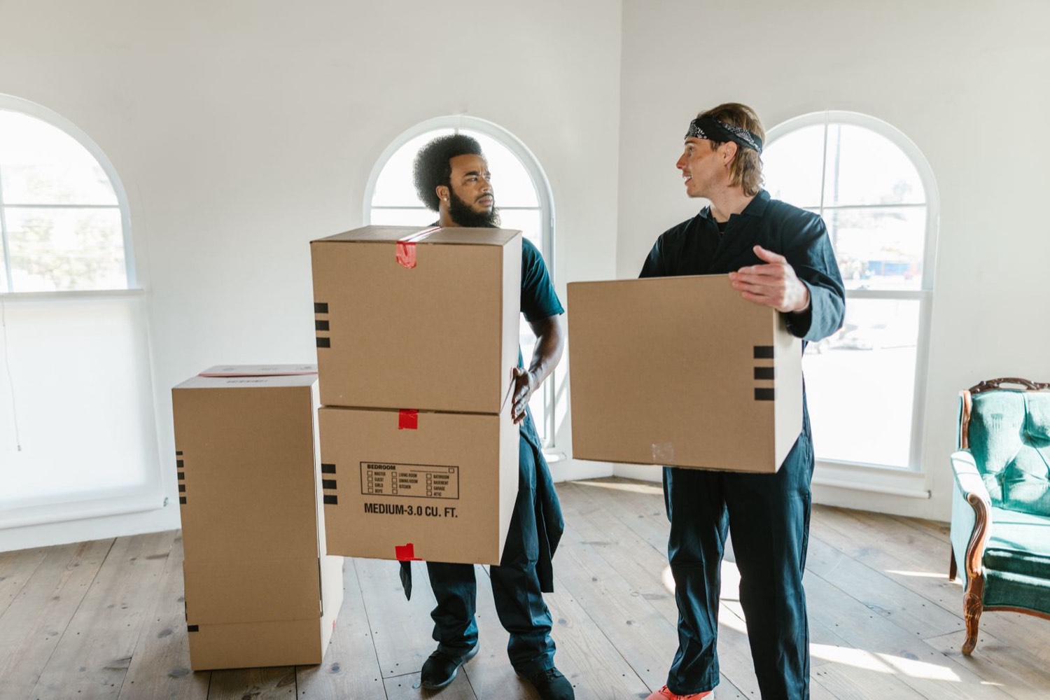 Two movers in dark uniforms carrying cardboard moving boxes through a bright, high-ceilinged room