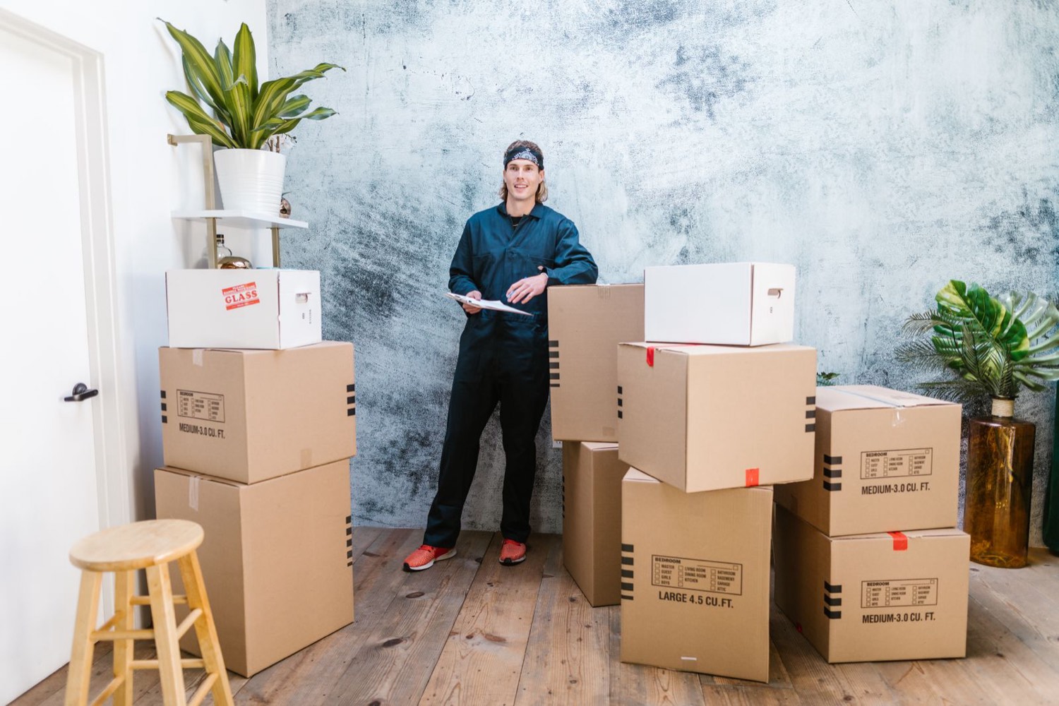 A mover in navy coveralls holding a clipboard while standing beside stacked, labeled moving boxes